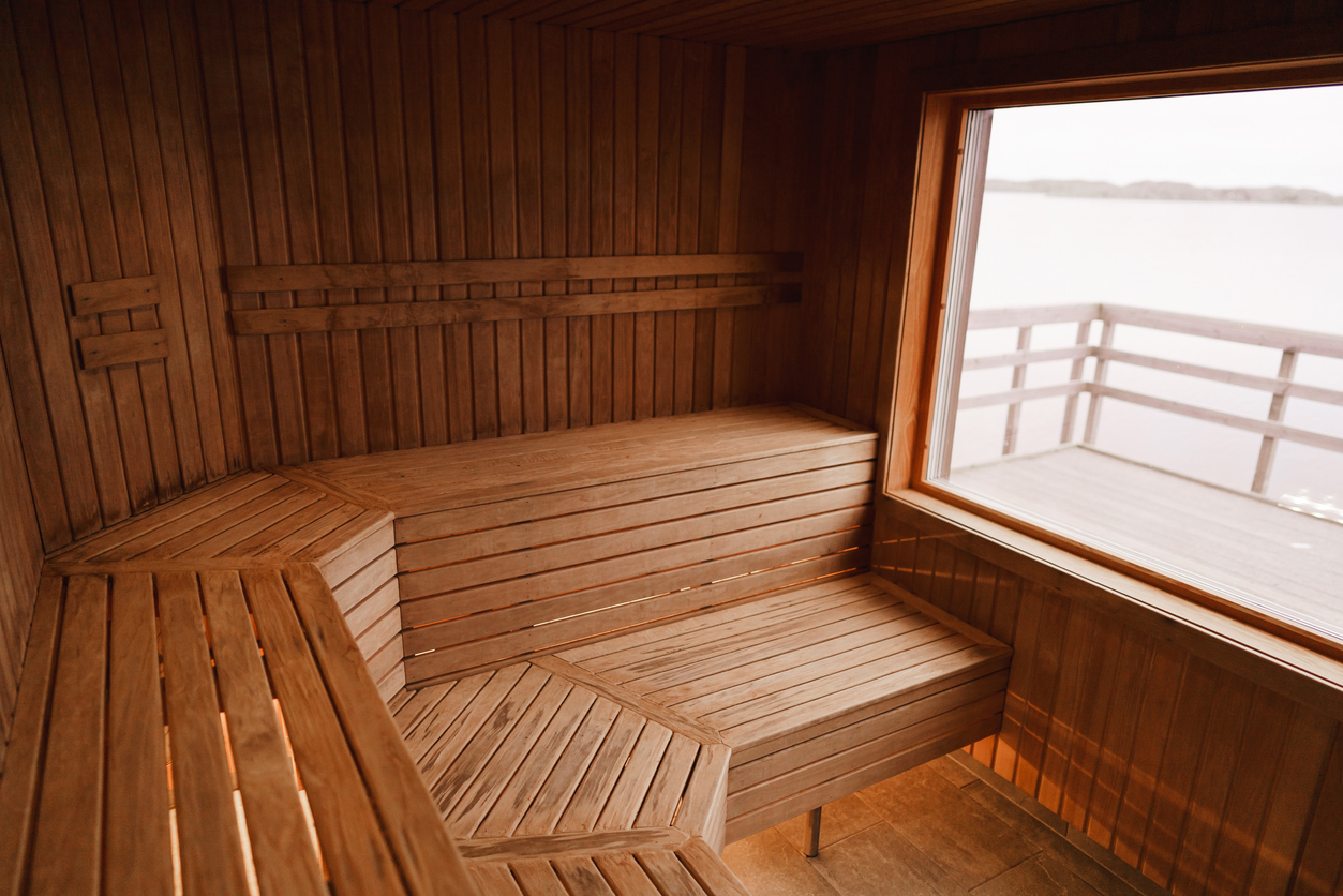 Photograph of the inside of an empty dry sauna with large windows. Walls and benches made of wood and a tile floor.